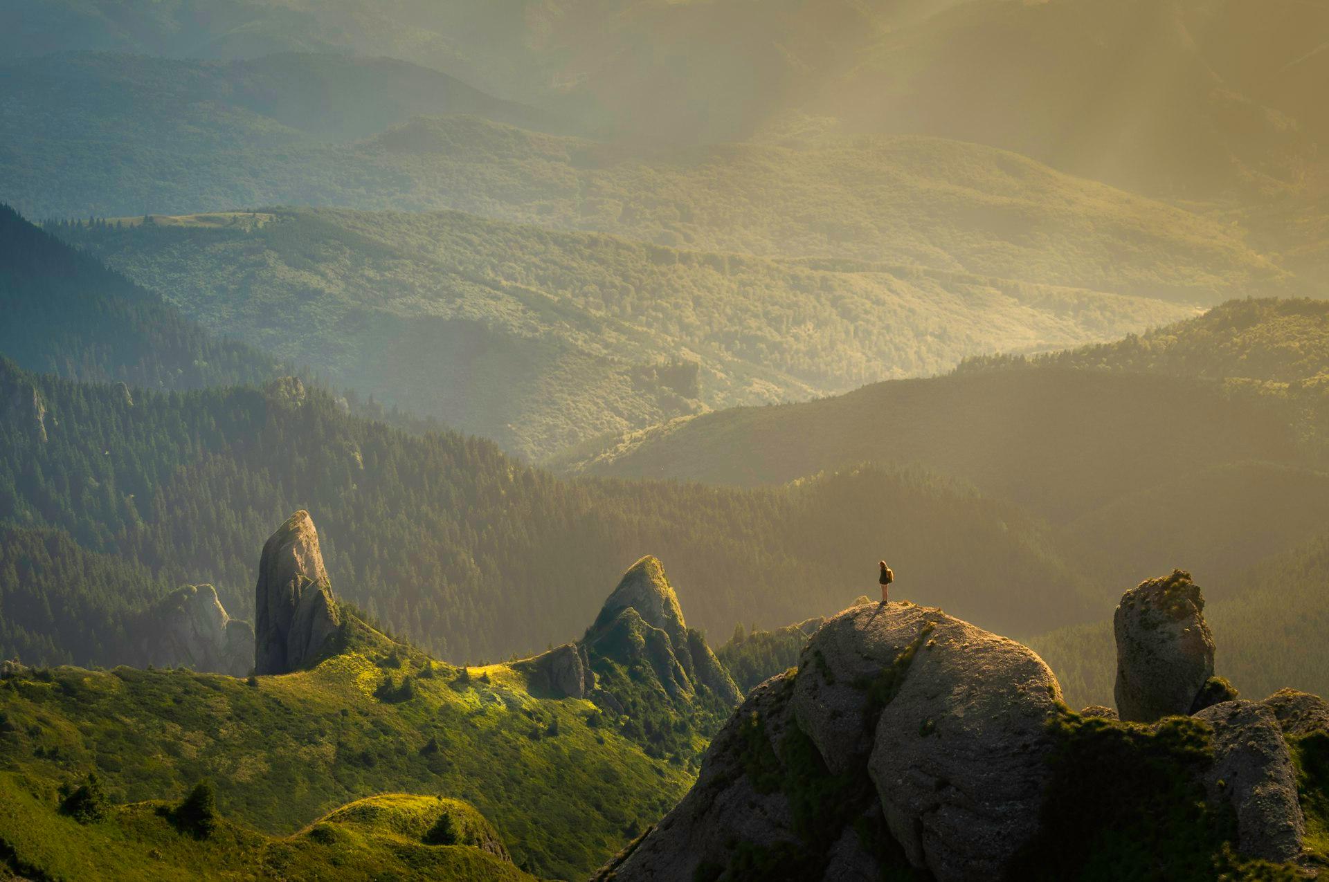 A lone figure standing on a rock outcrop above layered forested ridges in hazy golden light